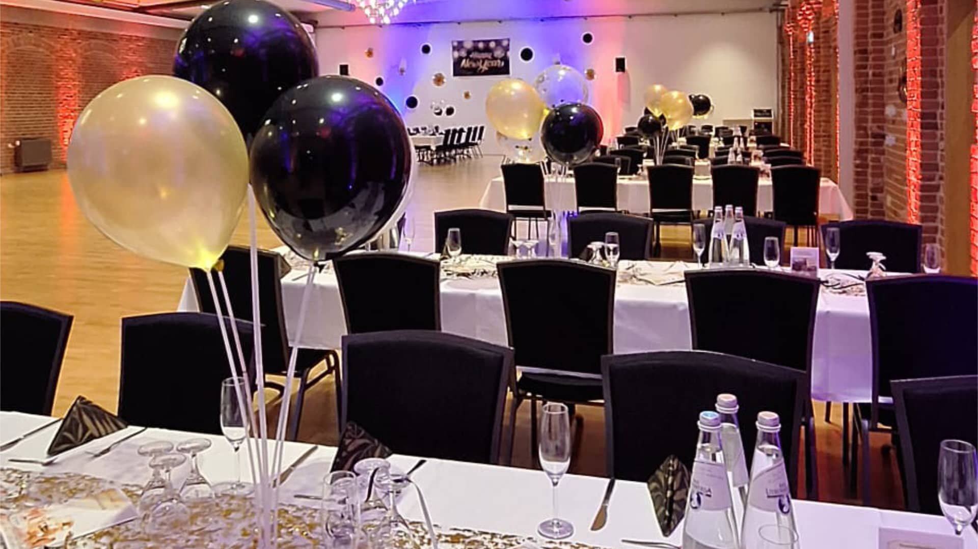 Dieses Bild zeigt eine festlich dekorierte Veranstaltungslocation, wahrscheinlich für eine Gala, ein formelles Abendessen oder eine Silvesterfeier. Die Farbpalette wird von elegantem Schwarz und Gold/Champagner dominiert, was auf einen gehobenen Anlass hindeutet. Im Vordergrund sind Tische mit weißen Tischdecken und dunklen Stühlen zu sehen, die für eine große Anzahl von Gästen gedeckt sind. Auffällige Dekorationselemente sind große, runde Ballons in Schwarz und Gold, die als Tischschmuck oder allgemeine Raumdekoration dienen. Die Konfetti- oder Glitzerdekoration auf dem Tisch verstärkt den festlichen Charakter.