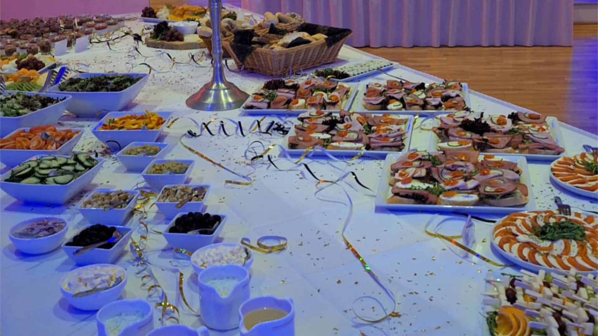 Dieses Bild zeigt ein reichhaltig gedecktes Buffet, wahrscheinlich bei einer formellen Veranstaltung, Feier oder einem Empfang. Die Beleuchtung ist gedämpft und von einem kühlen, bläulichen Licht dominiert, was eine elegante und festliche Atmosphäre schafft. Auf dem langen Tisch sind zahlreiche kleine Speisen angerichtet, was auf ein Catering-Arrangement hindeutet, das auf Fingerfood oder Häppchen spezialisiert ist. Man erkennt verschiedene Schalen mit Beilagen oder Salaten sowie Platten mit appetitlich arrangierten. Die Dekoration des Tisches ist dezent, mit goldenen oder glitzernden Akzenten, die sich über die weiße Tischdecke ziehen und die festliche Stimmung unterstreichen.
