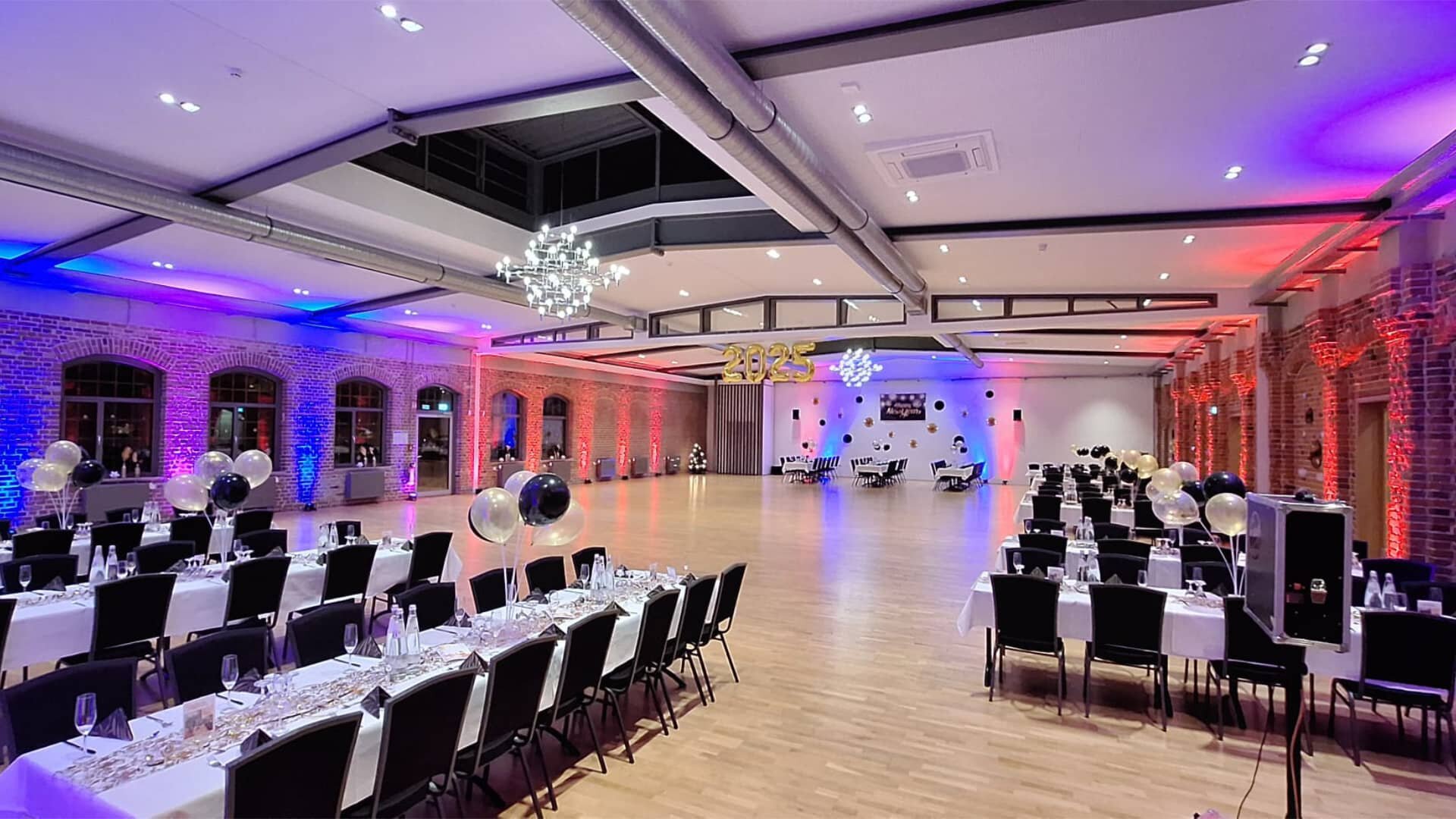 Dieses Bild zeigt einen großen, eleganten Veranstaltungssaal, der für eine Feier oder ein formelles Bankett vorbereitet ist. Die Atmosphäre wird durch eine dynamische Beleuchtung in Blau- und Rottönen an den Wänden und der Decke bestimmt, was auf eine festliche oder abendliche Veranstaltung hindeutet. Der Raum ist langgestreckt mit einem polierten Holzboden in der Mitte, der als Tanzfläche oder zentraler Bereich dient. Links und rechts sind lange Tische mit weißen Tischdecken und dunklen Stühlen aufgestellt, die auf eine große Anzahl von Gästen warten.