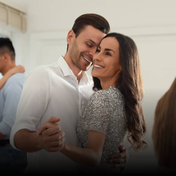 Das Bild zeigt ein Paar mittleren Alters, das eng umschlungen tanzt, wahrscheinlich auf einer Feier oder einem gesellschaftlichen Ereignis, was durch die festliche Kleidung der Frau (silbernes Paillettenoberteil) und das weiße Hemd des Mannes angedeutet wird. Die Beleuchtung ist hell und weich, was eine warme und intime Atmosphäre schafft. Die dominierende Emotion ist tiefe Zuneigung und Glück. Beide Personen lächeln strahlend und blicken sich mit großer Zärtlichkeit an. Die Körperhaltung – eng aneinander geschmiegt, Hände haltend – signalisiert Vertrautheit, Verbundenheit und Romantik. Es ist ein Moment der ungeteilten Aufmerksamkeit und Freude zwischen den beiden.