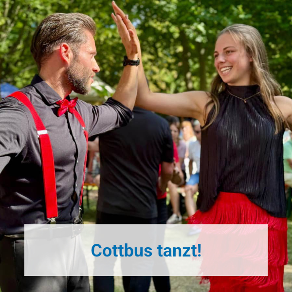 Das Bild fängt einen lebhaften Moment zwischen einem Mann und einer Frau ein, die wahrscheinlich an einem Tanzauftritt oder einer Tanzveranstaltung im Freien teilnehmen. Die Szene spielt sich an einem sonnigen Tag ab, erkennbar an dem hellen Licht und dem üppigen grünen Laub im Hintergrund, was auf eine Sommer- oder Frühlingsveranstaltung hindeutet. Der Mann trägt ein schwarzes Hemd, rote Hosenträger und eine rote Fliege, was auf einen klassischen, möglicherweise lateinamerikanischen oder Swing-Tanzstil hindeutet. Die Frau trägt ein schwarzes, plissiertes Oberteil und einen leuchtend roten Fransenrock, was ebenfalls stark auf einen Tanzstil wie Salsa, Tango oder Charleston schließen lässt.